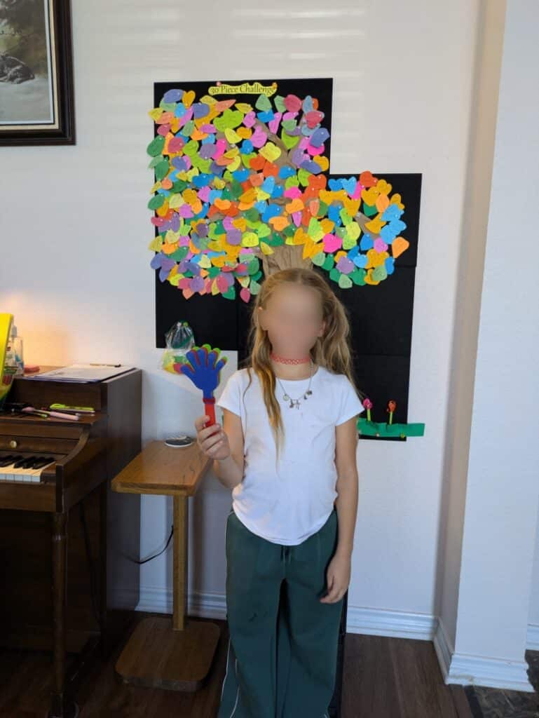Image of a smiling child holding an award and their chart of completed pieces in front of the studio decoration and incentive tree.