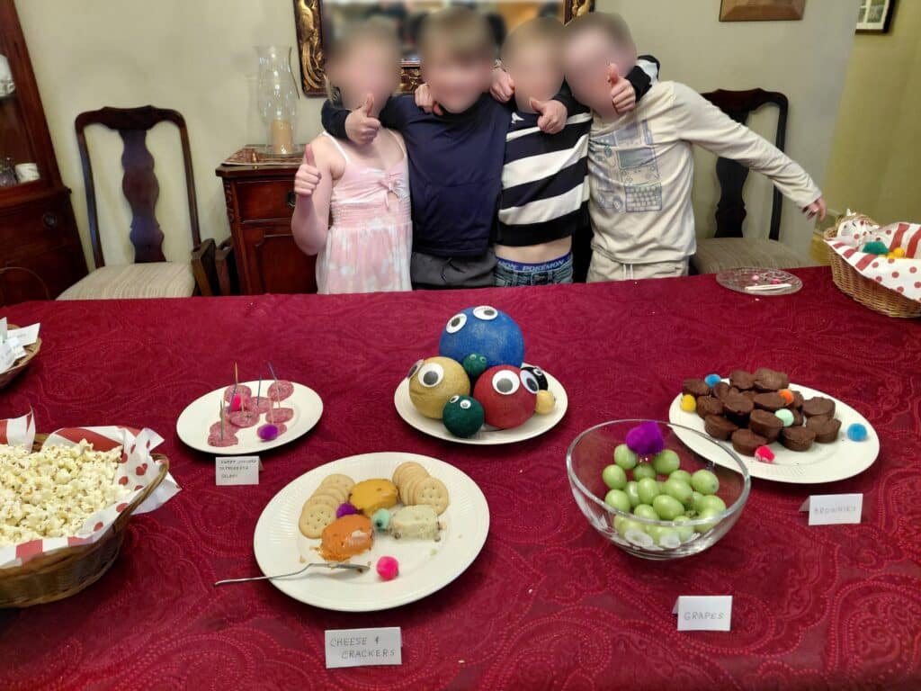 Little Fuzzies Party - 4 kids have gathered around a table of Little Fuzzies themed food. They're smiling and have their arms around each other for the photo, with thumbs up. Their faces are blurred for privacy.
